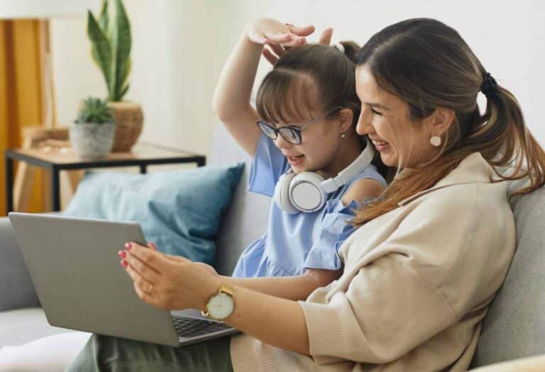 Mother and daughter sit on a sofa looking at a laptop screen, the girl wearing glasses with headphones around her neck and both smiling at the display.